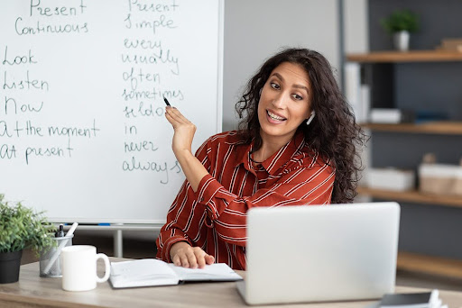 Young woman in a red striped shirt points at a whiteboard filled with notes beside an open laptop.